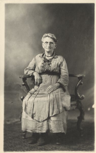 Full-length studio portrait of Margaret Pauhs (also spelled Paus), identified on back of the photograph as "Mrs. Werner Esser's mother," in front of a painted backdrop. She is seated in a chair and is wearing a dress with embroidery on the bodice. Pauhs was formerly Margaret Pick during her first marriage.