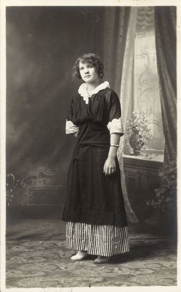 Full-length studio portrait of Mamie Bollenbeck Haack standing in front of a painted backdrop. She is wearing a dark dress with light collar and cuffs, and a striped underskirt. She has one hand behind her back.