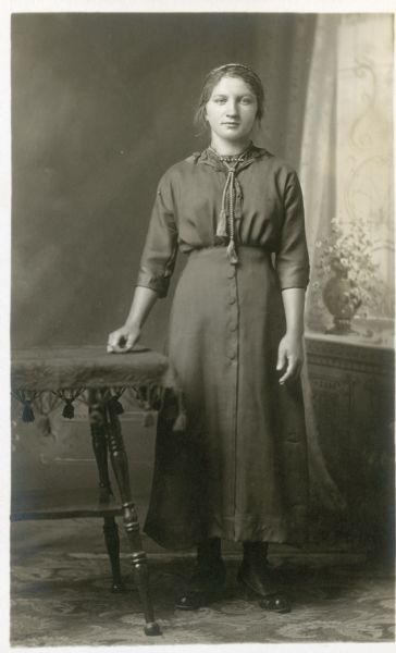 Full-length studio portrait of a standing woman wearing a dark dress in front of a painted backdrop. She is wearing a headband, and has a decorative cord around her neck, held in place with a bar pin. Her right hand rests on a table.
