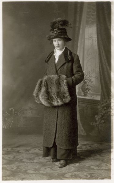Full-length studio portrait of Kathryn Dahman standing in front of a painted backdrop. She is wearing a long coat, a velvet hat with a feather plume, and is holding a fur muff.