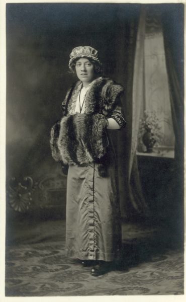 Full-length studio portrait of Frances Dahman, standing, wearing a dress in front of a painted backdrop. Over it she has a fur cape and muff, and is wearing a mob cap style hat.