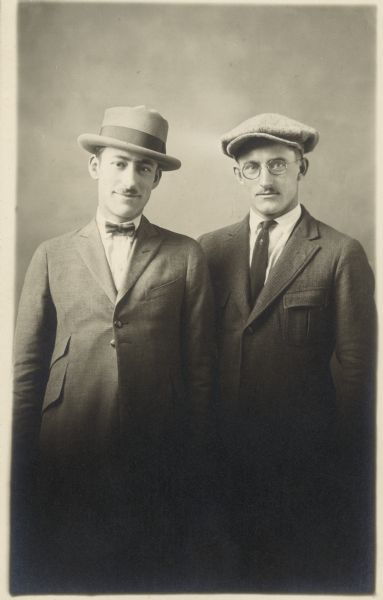 Three-quarter length studio portrait of Edward Faust and Frank Schulenburg. They are both wearing suits. One man is wearing a bow tie and hat, the other is wearing eyeglasses and a cap.