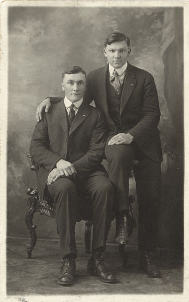 Full-length studio portrait of Melchoir Koch and Matt Oberembt in front of a painted backdrop. One man is sitting in a chair and the other is perched on the chair arm with his arm around the first man's shoulders. They are both wearing suits.