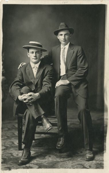 Full-length studio portrait of an unidentified man (left) and R. Melchior Endre in front of a painted backdrop. One man is sitting in a chair with his hands clasping his knee and Melchior is perched on the chair arm with his arm around the first man's shoulders. They are both wearing suits and hats.