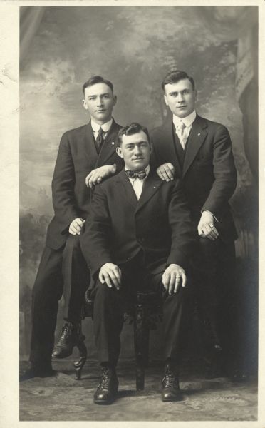 Full-length studio portrait of Mel Endres, Fred Statz and Robert Faust in front of a painted backdrop. The center man is seated in a chair, the men on either side are perched on the chair arms with a hand on the middle man's shoulders. All three men are wearing suits, and the seated man is wearing a bow tie.