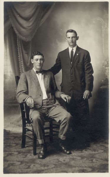 Full-length studio portrait of Max Esser and Robert Petersile in front of a painted backdrop. One man is seated and the other is standing on the right. They are both wearing suits.