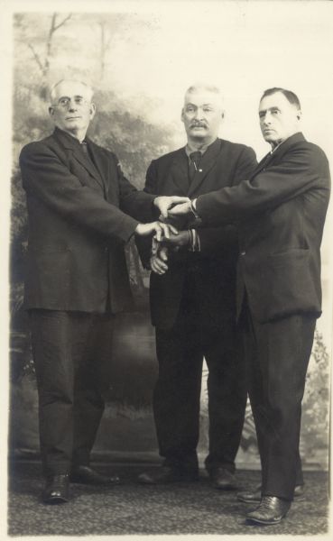Full-length studio portrait of Charles Meyer, Sr., James Gorman and an unidentified man, all wearing suits, posing in front of a painted backdrop. They have their hands stacked together in front of them.