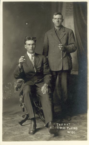 Full-length studio portrait of two men in front of a painted backdrop holding cigars. One man is sitting in a chair and the other stands to the right. The standing man is wearing a double-breasted suit jacket. Text in the lower right corner reads "Taken at Cross Plains, Wisc."