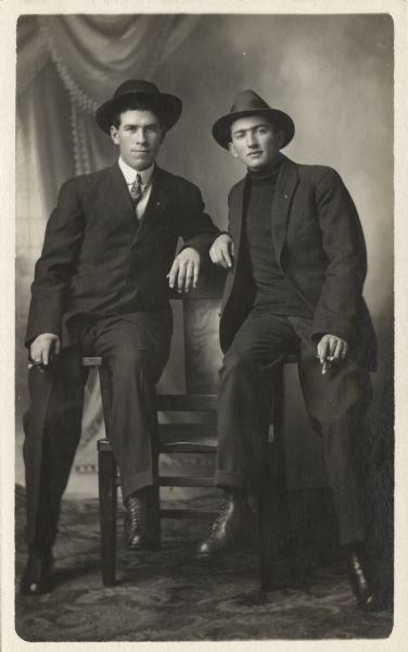 Full-length studio portrait of Mike Endres (right) and Carl Faust (left) in front of a painted backdrop. They are wearing suits and hats and are casually perched on the arms of a wooden chair and are both holding cigars.