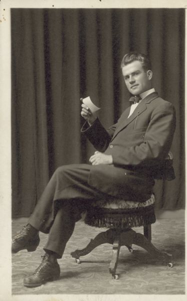 Full-length studio portrait of a man leaning back in a chair with his legs crossed, with a photograph is in his raised hand. He is wearing a suit with a bow tie and is gazing at the photographer. There is a curtain in the background.