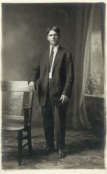 Full-length studio portrait of Robert Faust in front of a painted backdrop. He is standing with his right hand on a chair and is wearing a suit with the jacket unbuttoned.