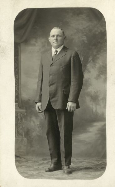 Full-length studio portrait of Albert Binger standing and wearing a suit and tie in front of a painted backdrop. The portrait has a white border with round corners.