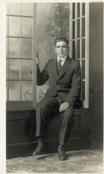 Full-length studio portrait of a man sitting on the sill of an open prop window in front of a painted backdrop. He is wearing a suit.