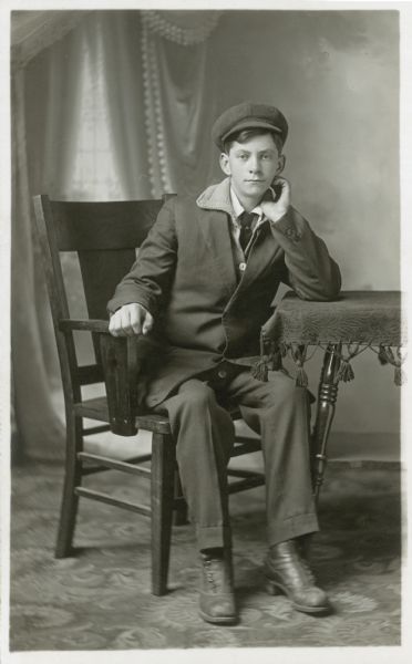 Full-length studio portrait of Hugo Faust sitting at a table in front of a painted backdrop. He is wearing a suit and cap and has one hand on the chair arm and other hand under his chin.
