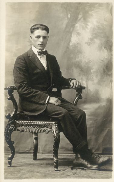 Full-length seated studio portrait in front of a painted backdrop of a man wearing a suit and a bow tie.