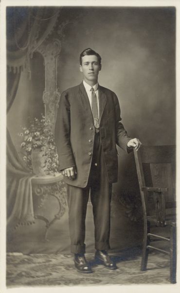 Full-length studio portrait of Frank Faust standing and wearing a suit with an unbuttoned jacket in front of a painted backdrop. His left hand is on the back of a chair.