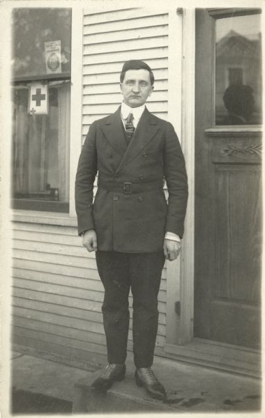 Portrait of Ollie Bordson standing on the stoop of a house. He is wearing a double breasted, belted coat over a suit. In the window to the left is a Red Cross sign and a United States Food Administration sign.