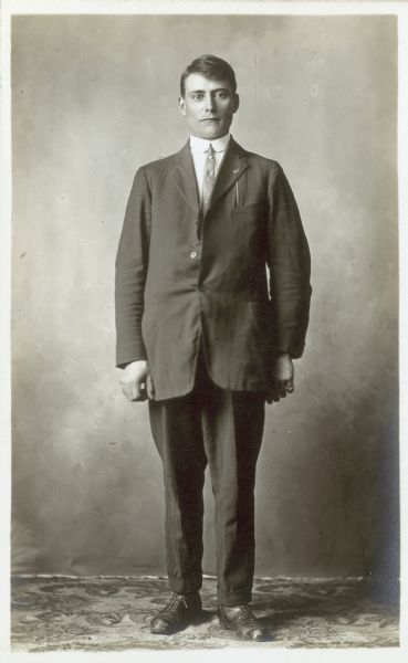 Full-length studio portrait of Theodore Falkenstein in front of a painted backdrop. He is wearing an ill-fitting suit.