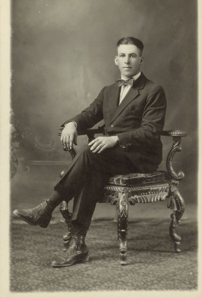 Full-length studio portrait of a man sitting in a chair with his legs crossed in front of a painted backdrop. He is wearing a suit with a bow tie.