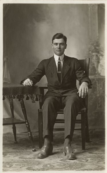 Full-length studio portrait of William Worringer in front of a painted backdrop. He is seated in a chair with his right forearm resting on a table.