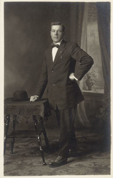 Full-length studio portrait of a man wearing a suit with a bow tie standing in front of a painted backdrop. He is resting his right hand on a table where his hat is placed, and has his left hand on his hip.