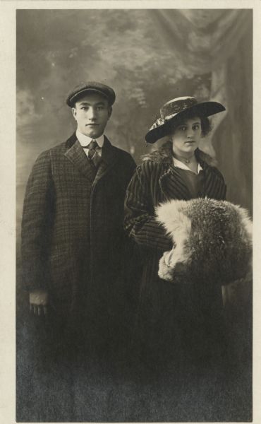 Photographic postcard of a three-quarter length studio portrait of Mrs. & Mrs. Otto Radke in front of a painted backdrop. They both wear hats and she holds a luxurious fur muff.