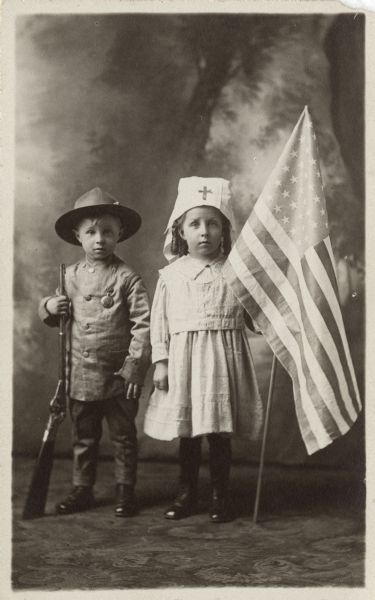 Full-length studio portrait of a girl dressed as a nurse and a boy dressed as a soldier standing in front of a painted backdrop. He holds a rifle and she is holding a United States flag.