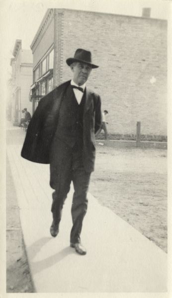 A man wearing a suit and hat is walking down the sidewalk. It is probably Frank Lloyd Wright. Several other men can be seen in the background.