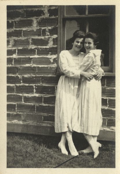 Salome Bollig and Regina Bollig Deans embrace and smile outdoors in front of a window in a brick wall. They are wearing light-colored dresses and lace-up boots.