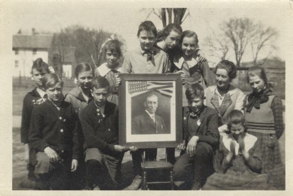 A civics class and teacher are displaying a portrait of President Woodrow Wilson. They are possibly gathered in the schoolyard.