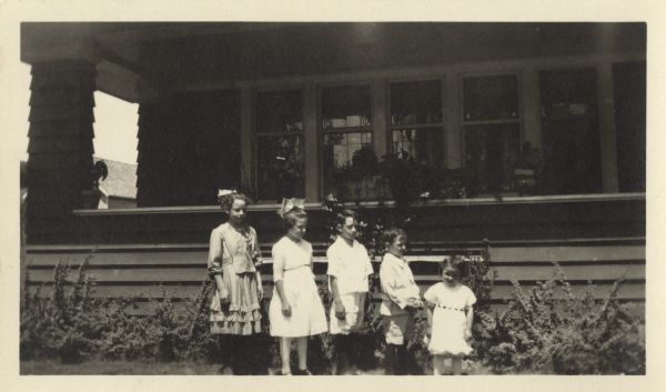 Five children pose in a row according to height in front of a porch. Their names are Theo Saeman Cavanaugh, Laura Saeman Gronenthal, Louis C. Saeman, Greg A. Saeman and Mary Angela Saeman.