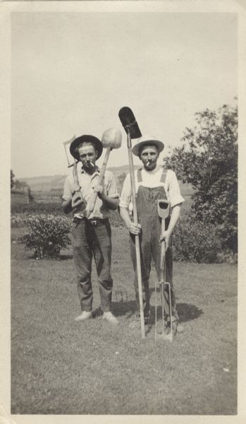 Two men in work clothes and hats pose in a yard with farm implements. Both of them are smoking cigarettes.