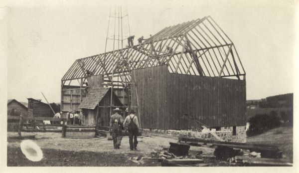 Outdoor scene of a crew of men building a barn. A smaller building is next to the tower structure for a windmill.