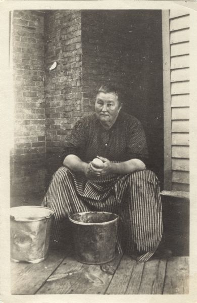 A seated woman peeling potatoes. Two buckets are on the floor in front of her and she is wearing a striped apron.