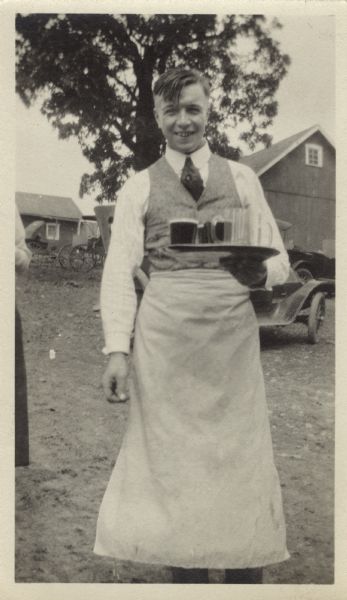 A smiling young man wearing an apron stands outdoors with a tray of drinks. Behind him are farm buildings, a buggy and an automobile.