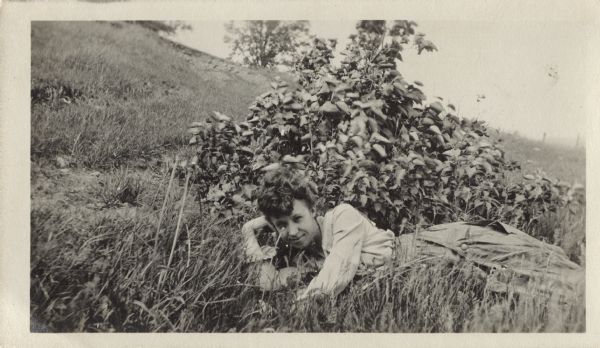A woman reclines on her side in the grass in front of a shrub. She is resting her head on her hand and is wearing a skirt and a blouse. In the background is a pasture and the slope of a hill.
