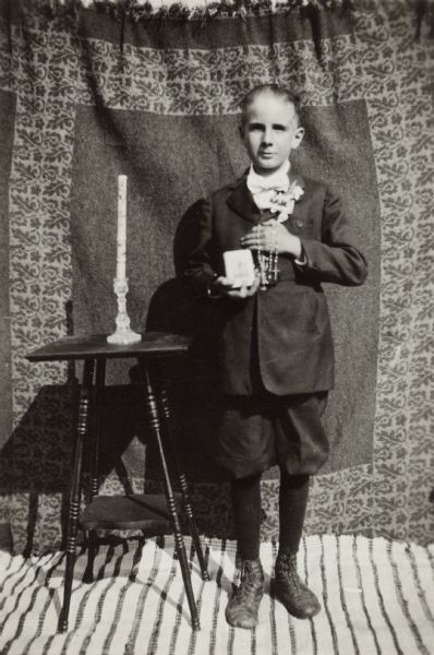 A boy poses for his first communion in front of a curtain near a table with a candle in a candle holder. He has a rosary wrapped around his hand and holds a catechism book in the other. He is wearing a jacket with a corsage pinned to it, knickerbockers (knickers), dark stockings and shoes.
