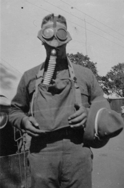 A man poses while wearing a gas mask. His hat is on his arm and he holds a pack in his hands with the strap around his neck. He appears to be wearing a military uniform. Trees and buildings are in the background.