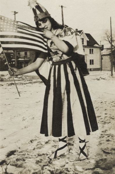 A woman wearing a striped patriotic costume poses outdoors with an American Flag. Her hat is topped with a metallic star, her ankles and shoes are wrapped with ribbons and her cape has stars on it. She is standing on the edge of a dirt road in a town.