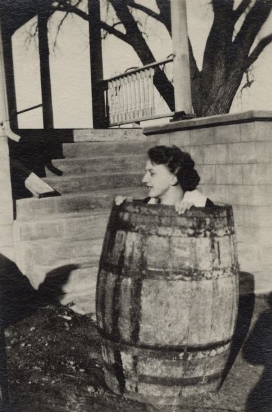 A woman crouches down inside a wooden barrel which is sitting on the ground near porch steps.