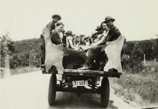 Rear view of group of men and women riding down a road in a wagon. The wagon has wooden benches (pews?) in it for passengers. Wooded hills can be seen in the background. The license plate is from Wisconsin, 27-568, WIS23.