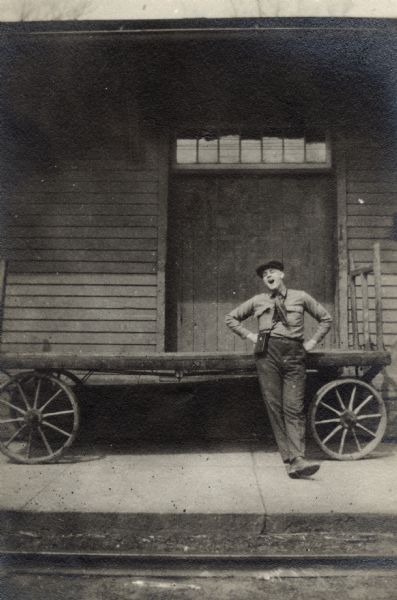 A man wearing a hat is leaning against a wagon with his hands on his hips. His feet are crossed and he appears to be speaking or singing. He has a case slung over his shoulder and a scarf around his neck. It is possible that he is a railroad worker. The wagon is parked on a platform, with railroad tracks in the foreground.