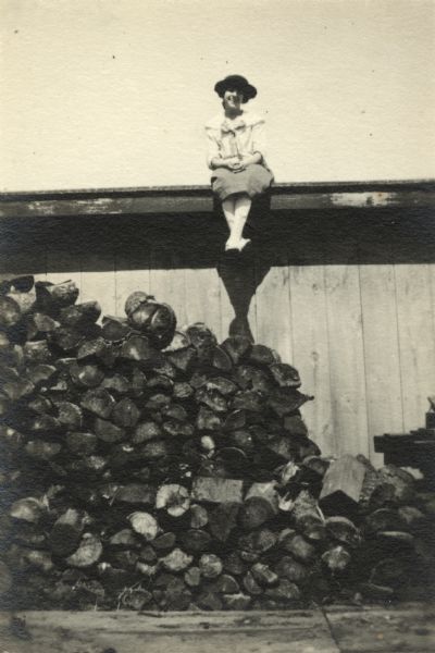 A woman perches on the edge of a roof over a wood pile. Her hands and feet are crossed and she is wearing a dress and hat.