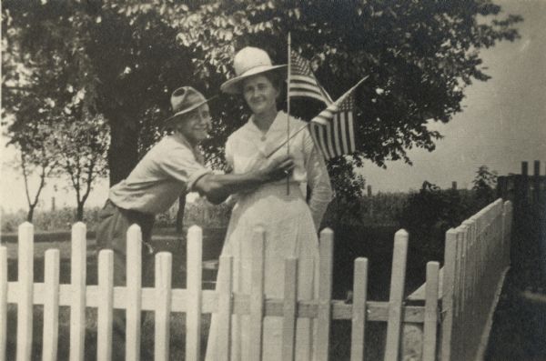 A man and a woman stand in a yard behind a white picket fence. The man is leaning towards her while holding an American flag. She is wearing a light-colored dress and hat, he is wearing a shirt, trousers and hat.