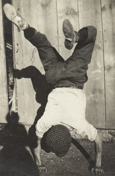 A man performs a handstand in front of a doorway, with his back to the camera. He is wearing a shirt, trousers, shoes and a plaid cap.