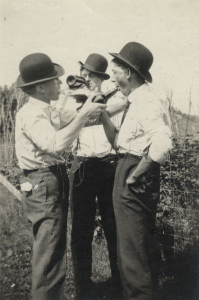 In a "gag" photograph, three men are drinking from bottles. The two men in the foreground are holding each others bottles. They appear to be standing next to a garden and are dressed in shirts, trousers, suspenders and bowler hats.