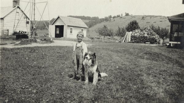 A boy stands in his yard with his dog by his side. He is wearing overalls and is barefoot. In the background (left to right) are farm buildings, semi-wooded hills, a woodpile and the porch of the farmhouse.