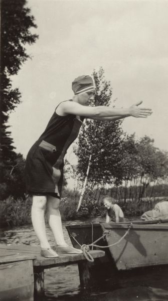 A woman dressed in a bathing suit, shoes and cap is about to dive off of a pier into the water. A rowboat is tethered on the right. Trees can be seen on the shoreline in the background. Another woman is in a boat hidden by the rowboat.