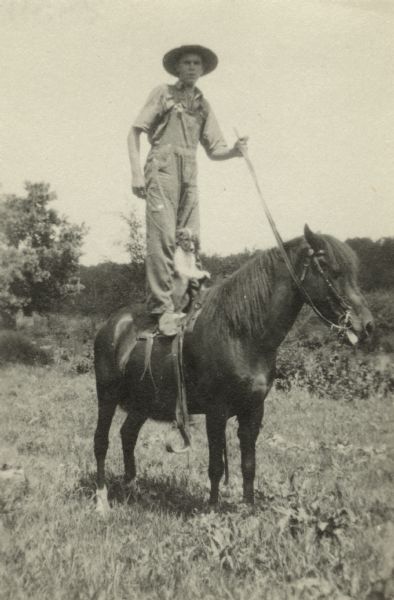 In a "gag" photograph, a teenager wearing overalls stands on the saddle of a pony, his dog tucked between his legs. He is wearing overalls and a hat. A field and woods appear in the background.
