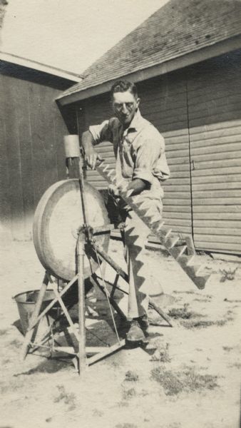 A man is seated at a grindstone sharpening a cutter bar. The bar was probably part of a piece of machinery used to cut hay or grain. He is dressed in work clothes. Behind him is a shed.